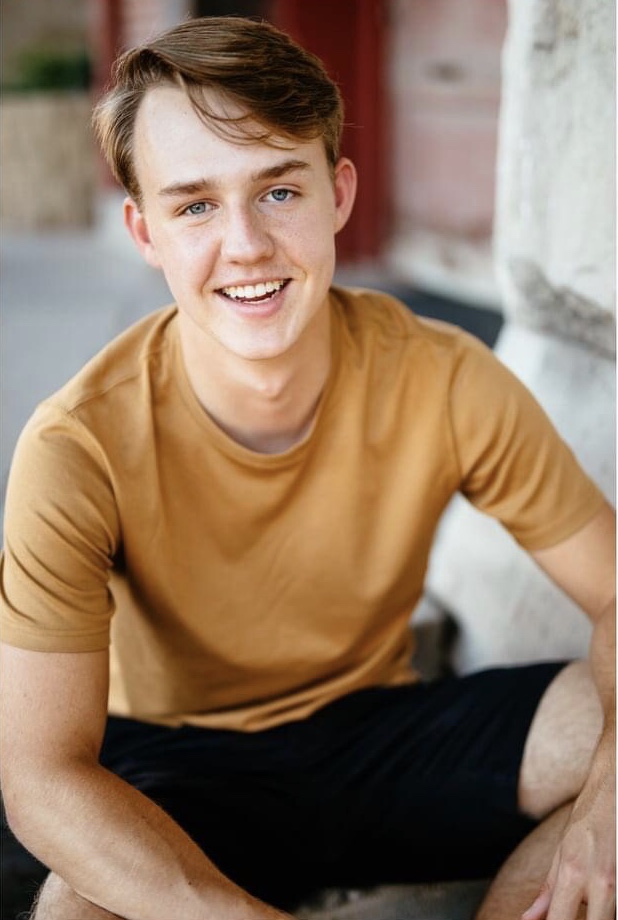 male high school student wearing a brown shirt and shorts sitting, posing for a senior photo