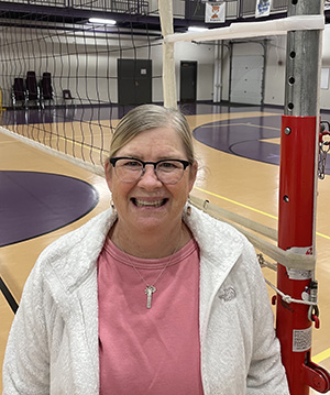 Woman smiling standing in front of a volleyball net wearing a pink shirt and white coat. 