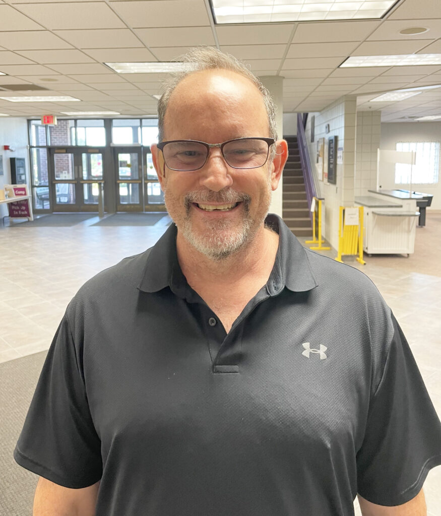 Man in a gray shirt smiling. He is wearing glasses.